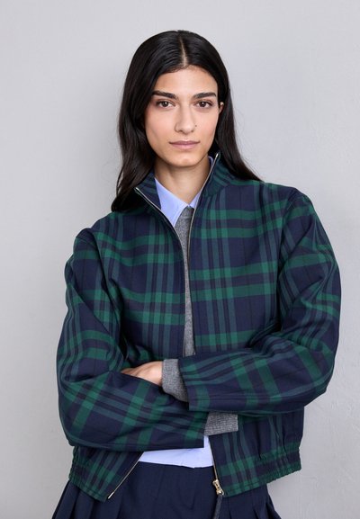 Giacca bomber a quadri blu navy e verde con zip frontale, collo rialzato e polsini a costine. Indossata sopra un maglione grigio e una camicia blu con colletto.