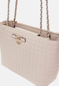 Bolso acolchado beige con herrajes dorados, que presenta asas de cadena y cierre de cremallera. Detalle del logo en la parte frontal y textura suave.