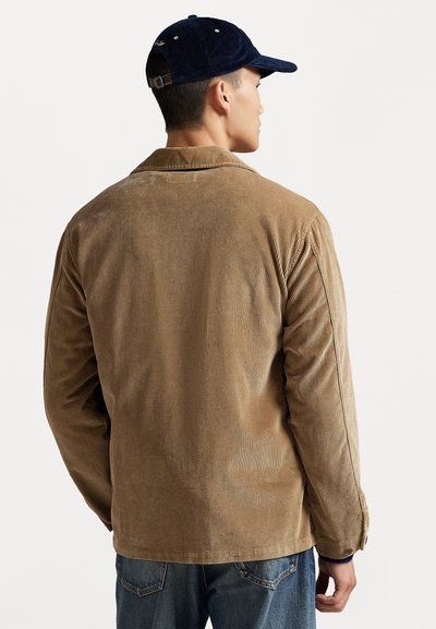 Brun corduroy-jakke med vertikale striper, med en klassisk krage og knappeledd, båret med en marineblå caps. Ledig passform.