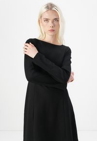 Svart langermet kjole med båthals, glatt tekstur og subtile sømmer. Tøyet har en avslappet passform og faller rett under knærne.