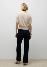 Beige Strickpullover mit kurzen Ärmeln, kombiniert mit dunkelblauen, hoch taillierten Jeans. Die Trägerin steht mit dem Rücken zur Kamera und zeigt das schlichte Design.