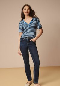 Randig blå och vit kortärmad topp med blå krage, kombinerad med mörk denimjeans. Modellen står framför en neutral bakgrund.