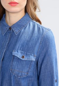 Nainen, jolla on päällään sininen farkkupaita, jossa on napitettavia rintataskuja ja kaulus, näkyvät huulet vaaleanpunaisessa huulipunassa, hiukset vaaleanruskeat.