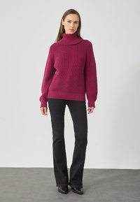 Sieviete, tērpta fuksijas krāsas apjomīgajā tērpi ar augstu apkakli, kombinēta ar melnajām platajām džinsu biksēm un melnām smailajām zābakiem pret neitrālu fonu.