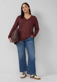 Burgunderfarbenes Langarm-V-Ausschnitt-Oberteil mit lockerem Schnitt, kombiniert mit weit geschnittenen blauen Jeans und sportlichen Sneakers, begleitet von einer braunen Schultertasche.