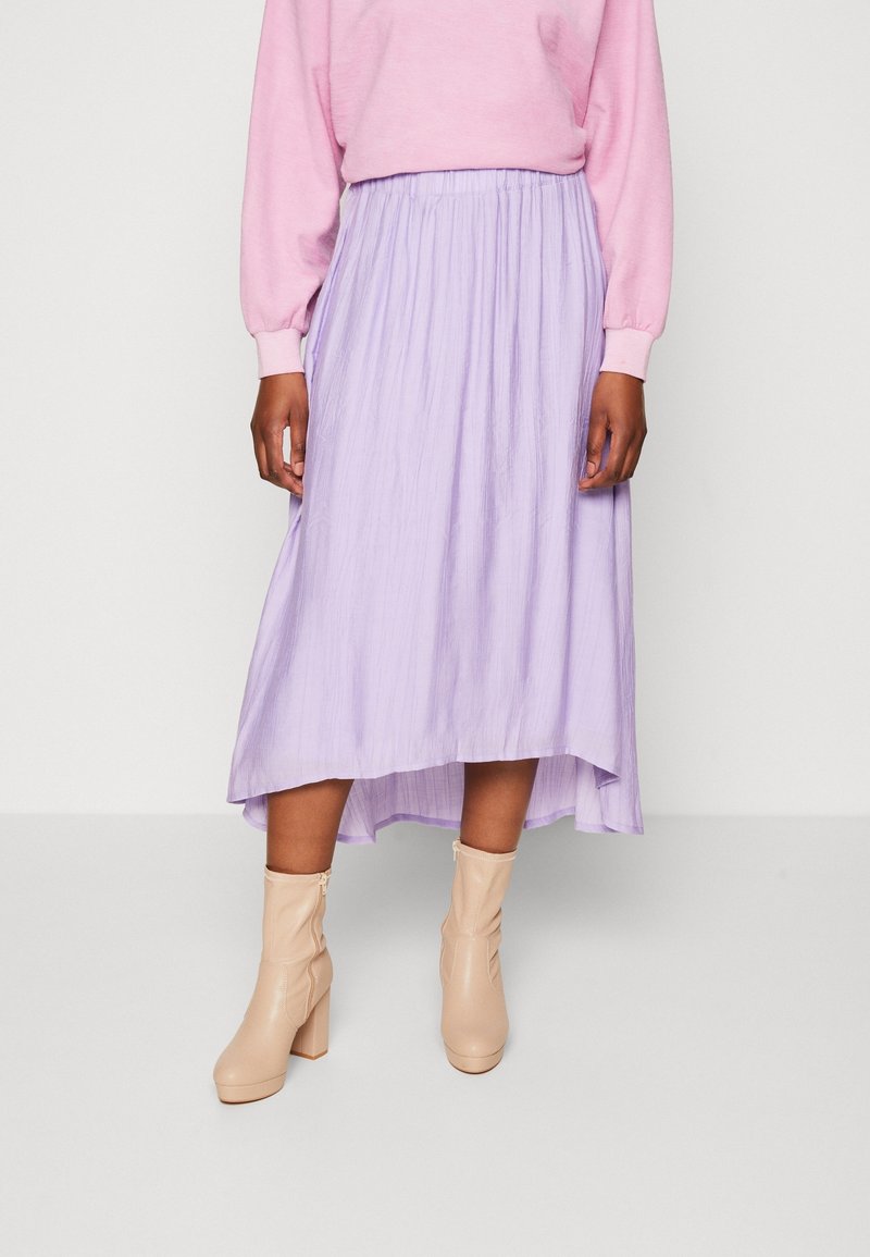 Femme portant une jupe midi violet clair, un pull rose à manches longues et des bottines beige, debout devant un fond blanc uni.