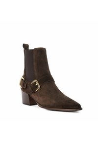 Bottine en daim marron avec un bout pointu, détail de boucle en or, panneaux latéraux élastiques et un talon empilé. Texture lisse et design minimaliste.