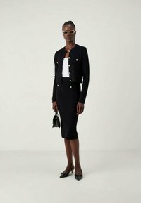 Veste en tricot noire avec des boutons dorés, haut blanc et jupe noire ajustée. Le modèle porte des lunettes de soleil et tient un petit sac à main noir.
