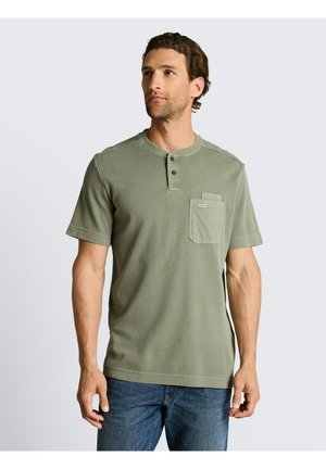 Mężczyzna w oliwkowej koszulce henley z krótkim rękawem i kieszenią na piersi oraz niebieskich dżinsach, stojący na prostym jasnym tle.