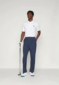 Hvit polo skjorte med et gult emblem, kombinert med marineblå bukser. Modellen holder en golfkølle og har på seg hvite golfsko med blå detaljer.