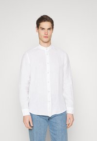Camisa de lino blanca con cuello tipo banda, cierre frontal de botones y mangas largas. Combinada con jeans de mezclilla azul claro. Diseño simple y texturizado.