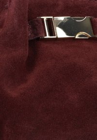 Bordeaux suède tas met een gladde textuur en een gouden metalen gespdetail op de band, dat een strakke uitstraling benadrukt.