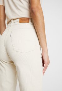 Beige kordfløyelsbukser med vertikal ribbet tekstur, med en lærmerkelapp og to baklommer. Subtil sømdetalj synlig.