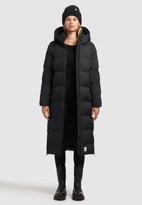 Piumino lungo nero con cappuccio, caratterizzato da un design a quadri imbottiti, aperture con zip e una toppa con logo all'orlo. Indossato con leggings neri e stivali.