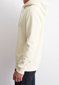 Felpa leggera beige con cappuccio, dotata di un cordino, texture morbida, polsini e orlo a costine. Presenta un dettaglio con logo vicino al bordo inferiore.