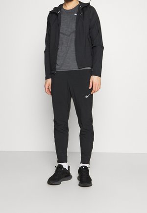 Completo sportivo nero che include una giacca leggera, una t-shirt grigia e pantaloni a gamba affusolata con un logo bianco. Sneakers nere con accenti bianchi.