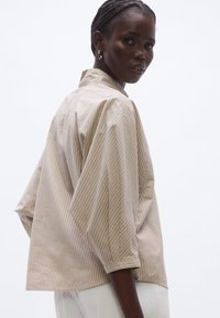 Cămașă bej cu dungi, guler înalt și design supradimensionat. Materialul este neted, cu mâneci lungi și largi și o croială lejeră.