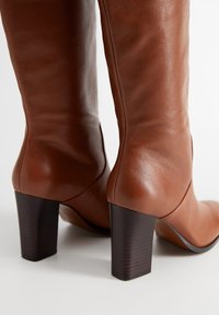 Bottes hautes en cuir marron jusqu'au genou, à texture lisse, talon carré et ajustement serré autour du mollet. Design minimaliste sans ornements visibles.