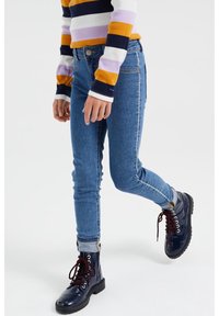 Magra blåjeans med en blekt tvätt, kombinerat med glänsande marinblå kängor med röda snören. T-shirten har flerfärgade horisontella ränder.