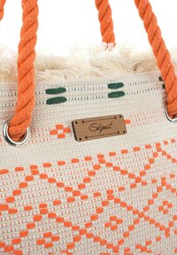 Bolso de mano tejido en crema con patrones geométricos naranjas. Presenta asas trenzadas naranjas, detalles con flecos y una etiqueta de marca de madera.