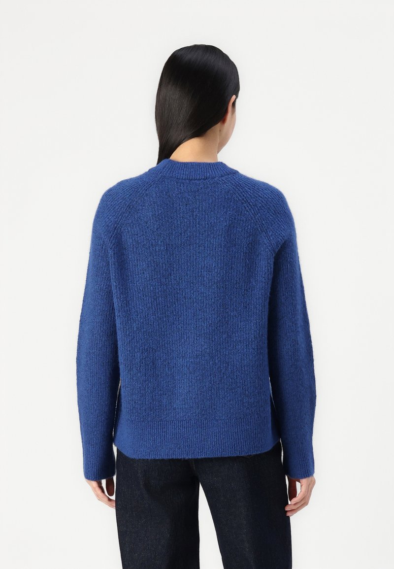 Persoon met lang zwart haar draagt een gestructureerde blauwe trui en donkere broek, gezien van achteren tegen een effen witte achtergrond.