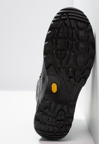 Schwarze Wanderschuhsohle mit robustem Profil, strukturiertem Gummi und einem auffälligen gelben Logo. Flaches, langlebiges Design für gute Traktion.