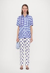 Camicia a maniche corte con bottoni a quadri blu chiaro e scuro, abbinata a pantaloni bianchi con grandi pois viola. Accessorio: borsa azzurro chiaro.
