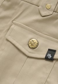 Beige tyg med en slät textur, som har en gyllene knapp med en ingraverad snöflinga och en svart etikett med "IBJ." i en accentuerad fickdesign.