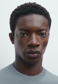 Portrét osoby s tmavou pletí a krátkými, texturovanými černými vlasy, oblečené v světle šedém tričku. Pozadí je neutrální a jemné.