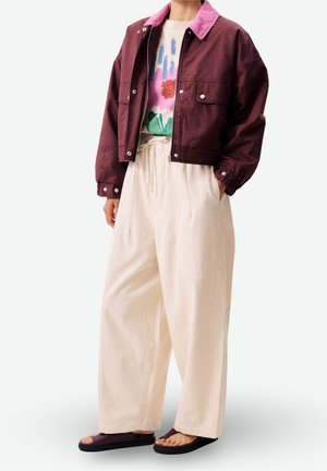 Personne portant une veste grenat par-dessus un t-shirt fleuri, un pantalon crème ample et des sandales ouvertes aux orteils violet foncé, debout les mains dans les poches.