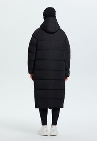 Abrigo puffer largo negro con capucha, con secciones acolchadas y un corte relajado. Combinado con leggins negros y zapatillas blancas.