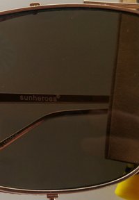 Sunglasses med mörka linser och en ram i guldmetall. Bågarna har varumärket "sunheroes" i vit text.