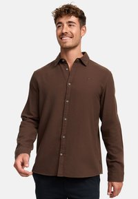 Camisa marrón de manga larga hecha de una tela suave, presenta un cuello clásico, cierre frontal con botones y un pequeño logo en el pecho.