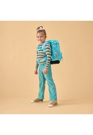 Lächendes Mädchen, das mit einem großen blauen Rucksack steht, trägt ein gestreiftes Langarmshirt, türkise Hose und grüne Turnschuhe vor beigem Hintergrund.