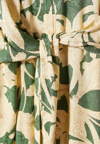 Tissu vert et crème avec un grand motif floral botanique noué, montrant la texture et les plis.