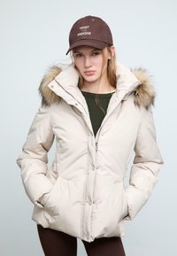 Giubbotto puffo beige con colletto alto e bordino in pelliccia, con chiusure a zip e a pressione, tasche laterali e una texture liscia. Cappello marrone con testo.