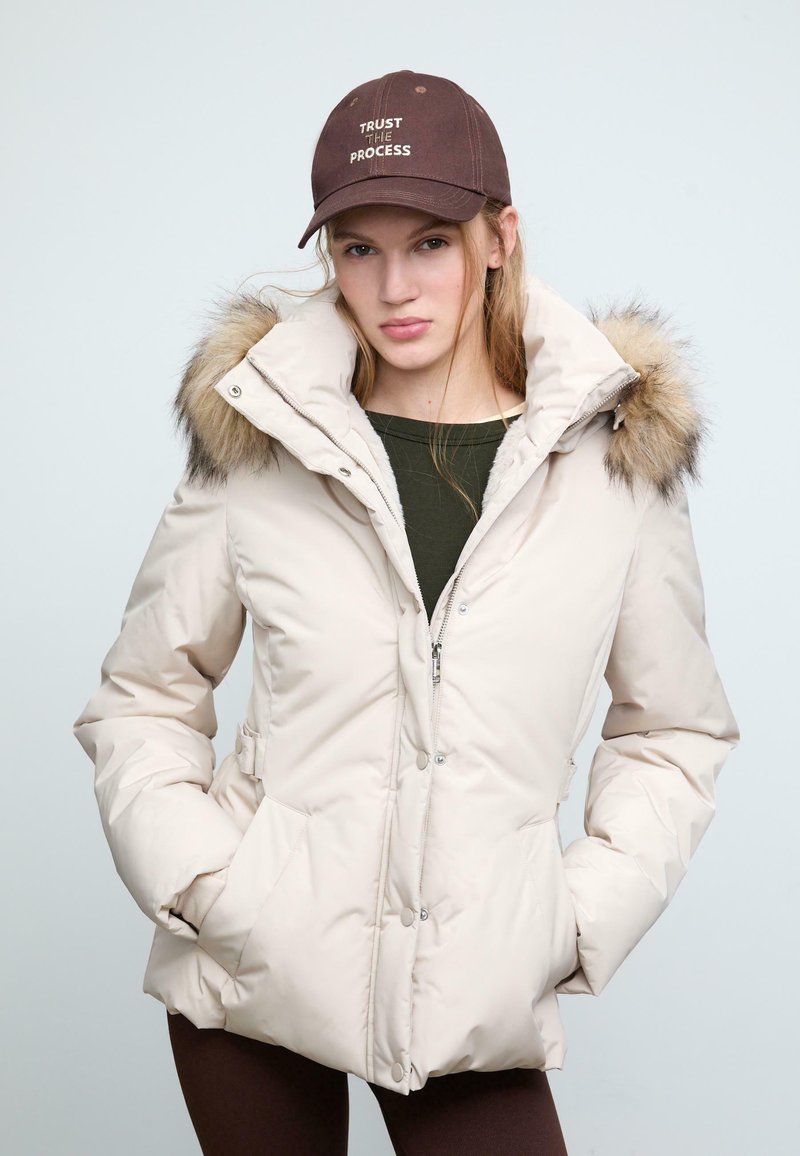 Giubbotto puffo beige con colletto alto e bordino in pelliccia, con chiusure a zip e a pressione, tasche laterali e una texture liscia. Cappello marrone con testo.