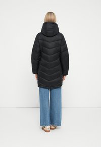 Doudoune noire avec capuche, présentant un design en chevrons. Associée à un jean en denim bleu à jambes larges et des chaussures de couleur claire.