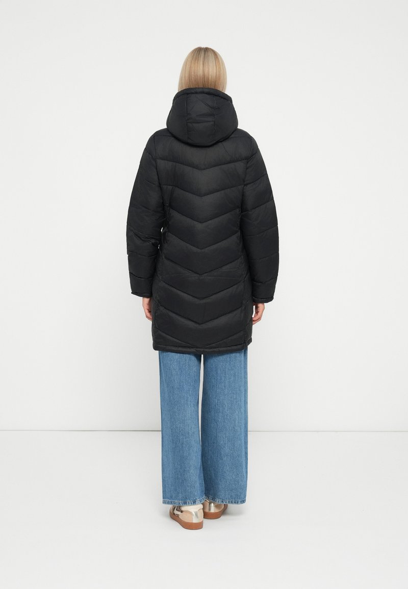 Schwarze Steppjacke mit Kapuze, gekennzeichnet durch ein Chevron-Steppdesign. Kombiniert mit weit geschnittenen blauen Jeans und hellen Schuhen.