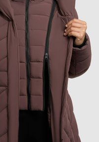 Chaqueta acolchada tipo puffer en marrón oscuro con cremalleras negras. Presenta un frontal con cremallera y capucha, exhibiendo un tejido acolchado y texturizado.
