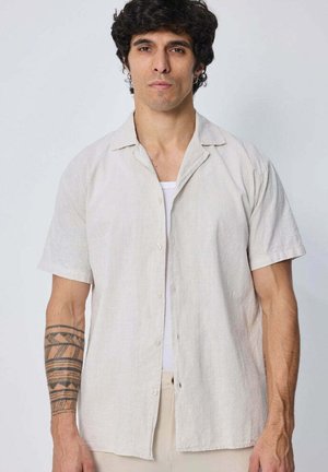 Homme aux cheveux foncés et bouclés porte une chemise claire à manches courtes par-dessus un haut blanc, avec un tatouage géométrique sur l'avant-bras gauche.