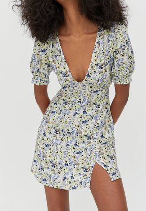 Femme portant une robe fleurie à manches courtes avec un décolleté en V profond et une fente sur la cuisse, debout devant un fond clair uni.