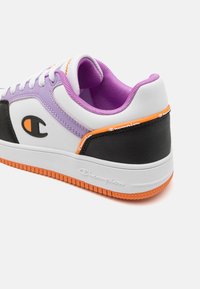 Białe sneakersy z czarnymi i purpurowymi akcentami, charakteryzujące się teksturowaną powierzchnią oraz gumową podeszwą z pomarańczowymi detalami. Widoczne logo Champion.