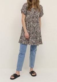 Czarna i beżowa tunika w wzory z krótkimi rękawami, połączona z jasnoniebieskimi dżinsami i czarnymi klapkami z detalem w postaci złotej klamry.