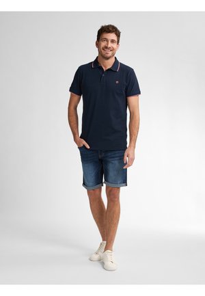 Camisa polo navy con acento rojo en el cuello, combinada con pantalones cortos de denim arremangados y zapatos blancos de cordones. El modelo está de pie de forma casual, con las manos en los bolsillos.