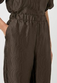 Jumpsuit marrón, texturizado con un patrón de olas, con cinturilla elástica y bolsillos laterales. Fabricado en un tejido suave y brillante.