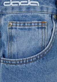 Svijetloplava denim tkanina ima stražnji džep s žutim šavovima i ukrasnim srebrnim gumbom. Na vrhu se nalazi bijeli logo.