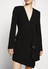 Robe blazer noire avec un profond décolleté en V, manches longues, boutons croisés, poches latérales et un détail drapé à l'avant. Texture de tissu lisse.