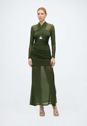 Femme portant une longue robe transparente vert olive avec un haut croisé, un détail trou de serrure, une taille froncée et des talons ouverts dorés, debout sur fond blanc.