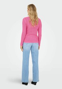 Femme aux longs cheveux blonds portant un pull en tricot rose, un jean à jambes larges bleu clair et des chaussures noires à talons, debout de dos.